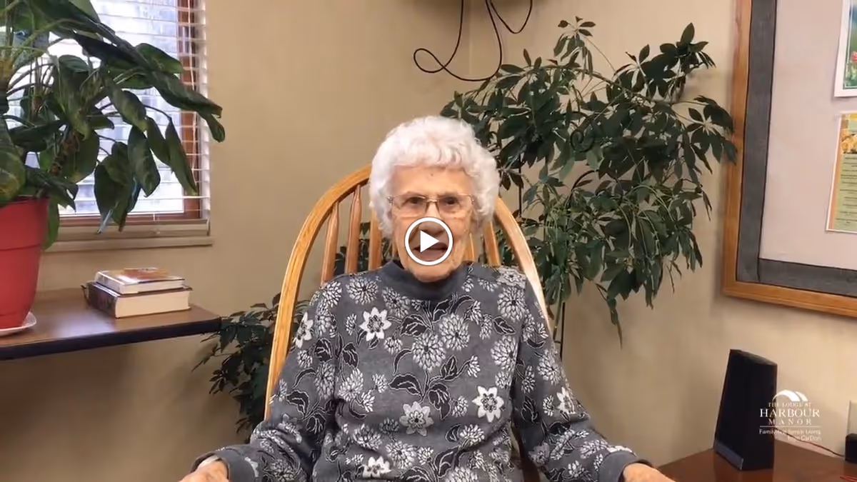An elderly woman with white hair and glasses sits in a wooden rocking chair in a cozy room with beige walls. Behind her is a large green plant, a window with blinds, and a table with books and a red plant pot. On the right side, there is a framed bulletin board and a small black speaker on a wooden surface.