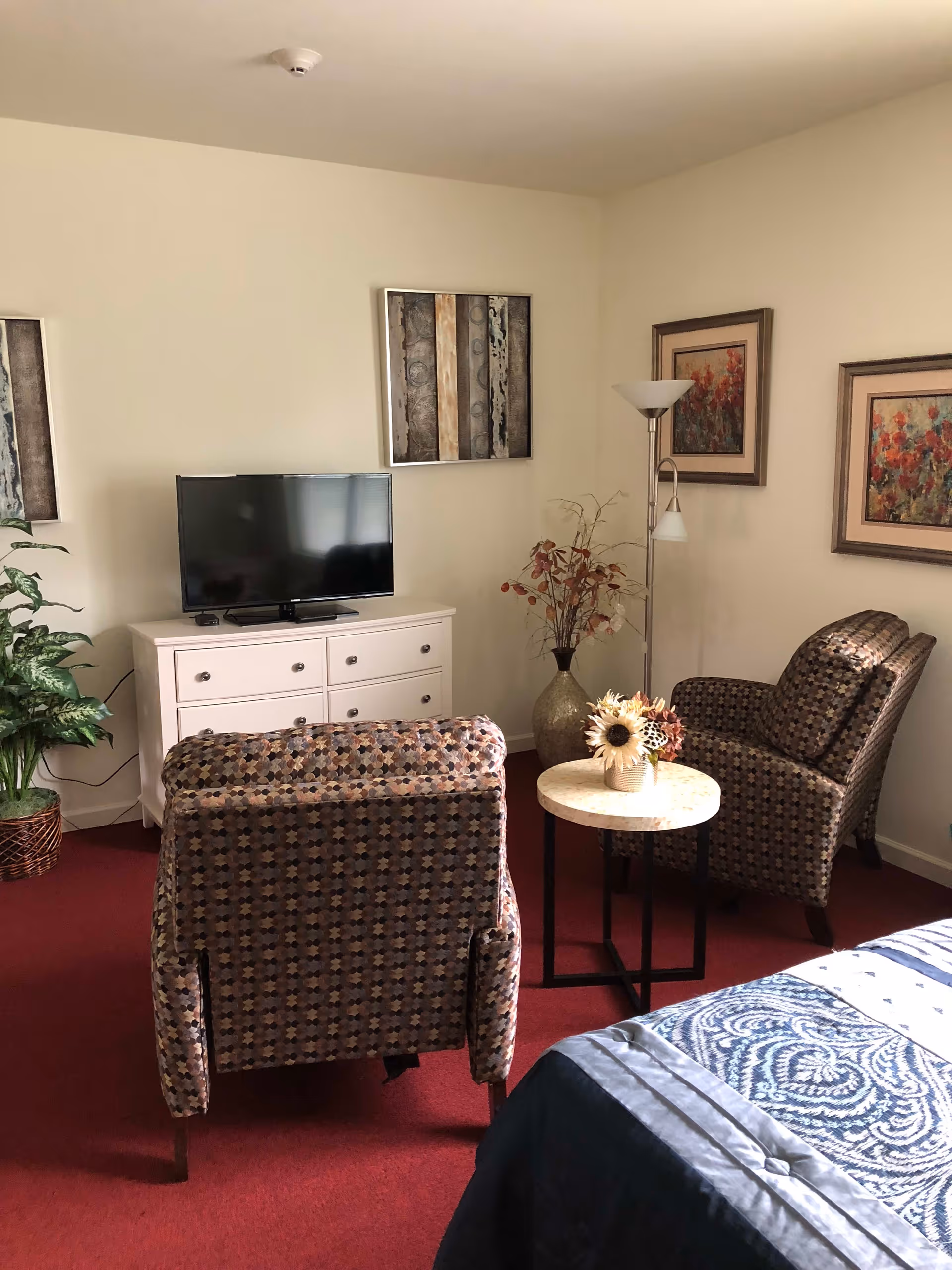 A cozy living area with two patterned armchairs facing a flat-screen TV on a white dresser. There is a small round table with a vase of flowers between the chairs, a floor lamp, and framed artwork on the walls. A bed with a blue patterned blanket is partially visible in the foreground, and a potted plant is in the corner.