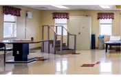 A spacious room with polished floors featuring physical therapy equipment including parallel bars and a set of stairs for rehabilitation exercises. The room has three windows with maroon valances and a bench along the right wall.