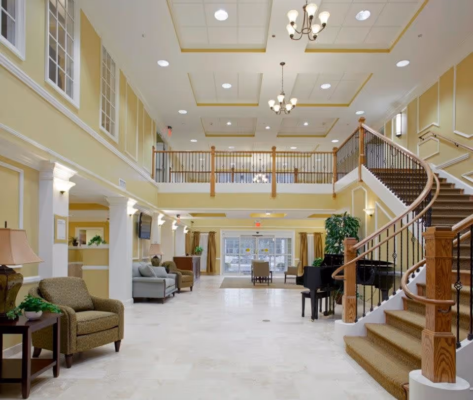 Spacious and well-lit interior lobby area of a senior living facility with a grand staircase on the right, comfortable seating areas with armchairs and sofas on the left, a black grand piano near the staircase, and large windows and glass doors at the far end letting in natural light. The walls are painted light yellow with white trim, and the ceiling features recessed lighting and chandeliers.