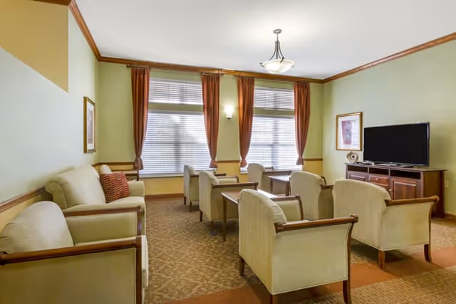 A well-lit living room with beige armchairs and sofas arranged around a central area. There are three large windows with brown curtains letting in natural light. A wooden cabinet holds a flat-screen TV on the right side of the room, and a framed picture hangs on the wall above it. The walls are painted light green with wooden trim, and a ceiling light fixture is centered above.