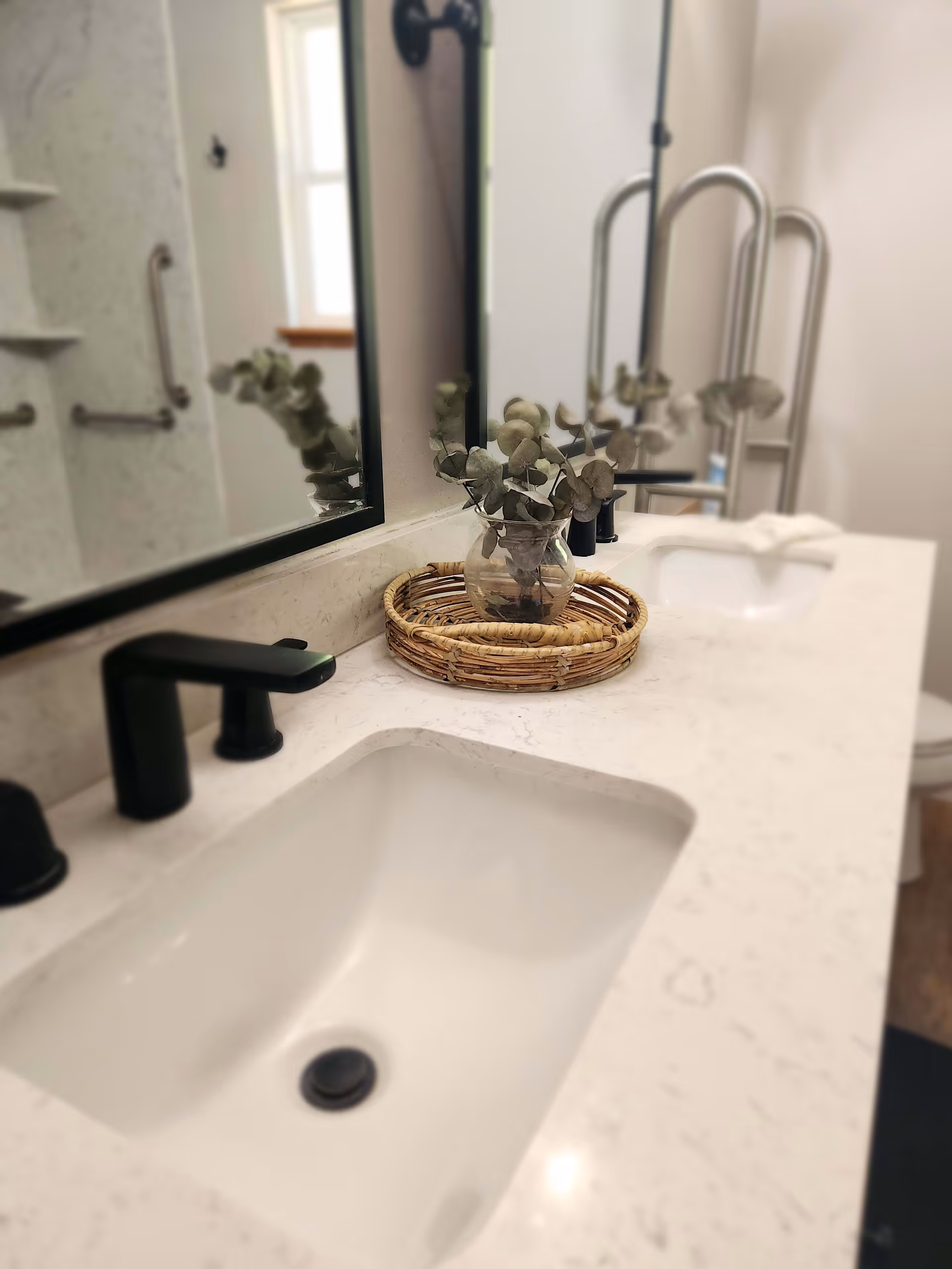 A bathroom countertop with two white sinks and black faucets. A round wicker tray with a glass vase holding eucalyptus branches is placed between the sinks. A large mirror is mounted on the wall above the countertop, reflecting part of the bathroom including grab bars and a window.