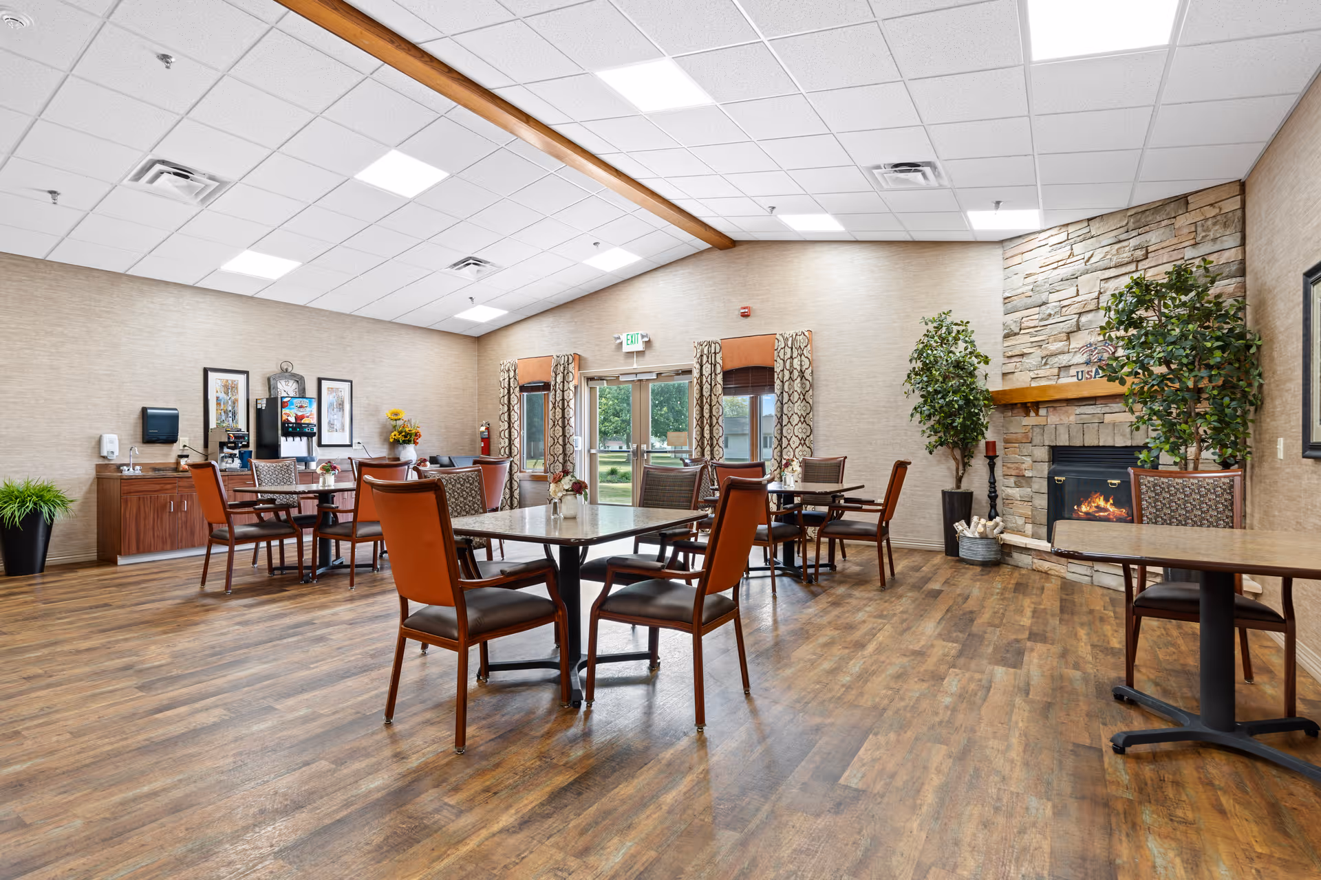 A spacious dining room with several tables and chairs arranged on a wooden floor. The room features a stone fireplace with a fire burning, two large potted plants on either side, and large windows with patterned curtains letting in natural light. There is a beverage station with a coffee machine and framed artwork on the wall. The ceiling has recessed lighting and a wooden beam running across it.