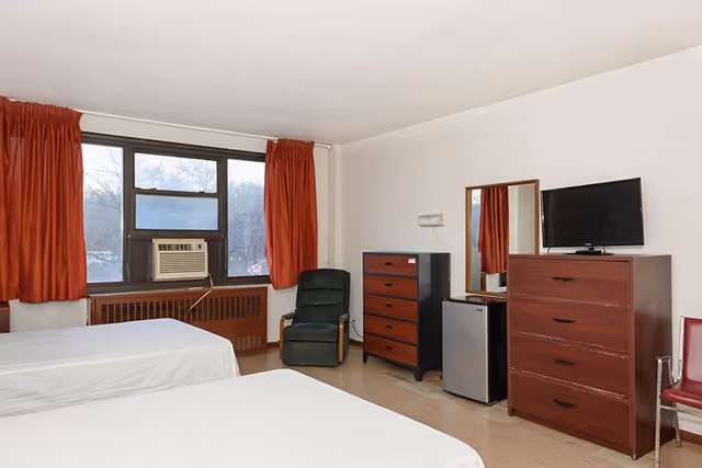 Simple shared bedroom with two beds, red curtains, a window air conditioner, a green armchair, and dressers with a TV.