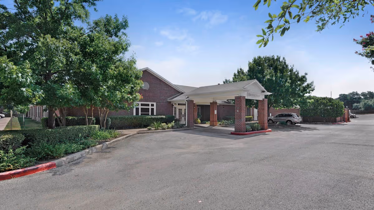 Front entrance of a brick senior living facility with a covered porte-cochere, trees, and a parking lot.