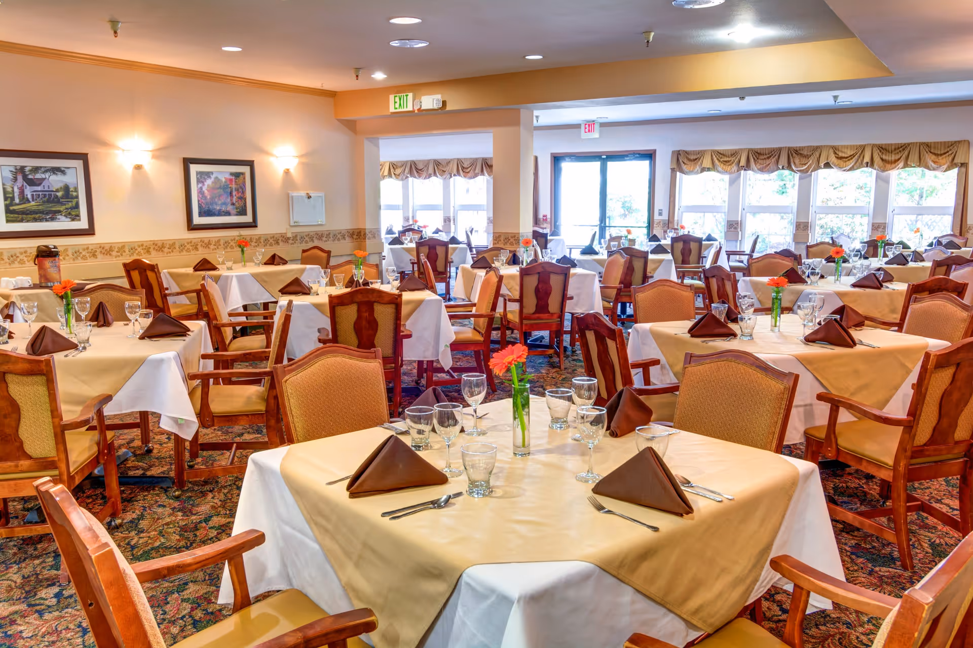 A bright dining room with multiple tables covered in white and beige tablecloths, each set with brown folded napkins, glassware, and silverware. The room has wooden chairs with cushioned seats and backs, floral carpet, framed artwork on the walls, and large windows letting in natural light.
