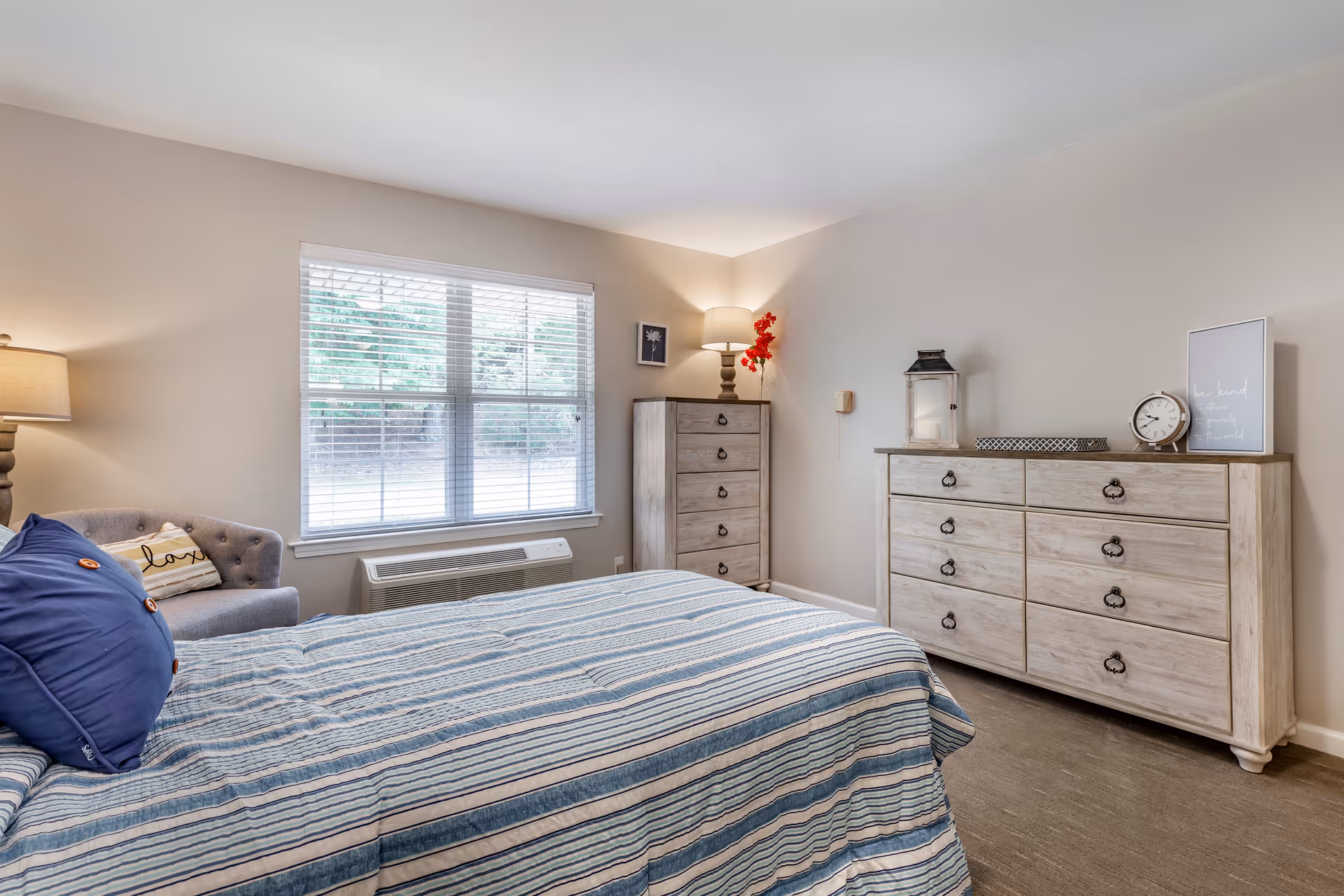 A bedroom with a bed covered in a blue and white striped comforter and blue pillows. There is a gray armchair with a pillow next to a window with white blinds. Against the wall are two light wood dressers, one tall and one wide, with a lamp, a clock, a lantern, and decorative items on top. The walls are painted light beige and the floor is carpeted.