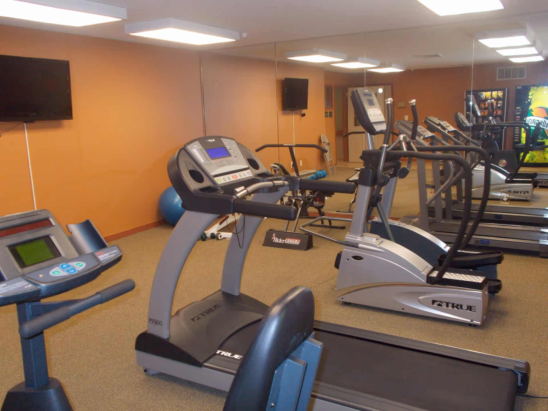 Indoor fitness room with exercise equipment including treadmills, elliptical machines, and a stationary bike. The room has orange walls, a large mirror on one side, a wall-mounted TV, and vending machines in the background.