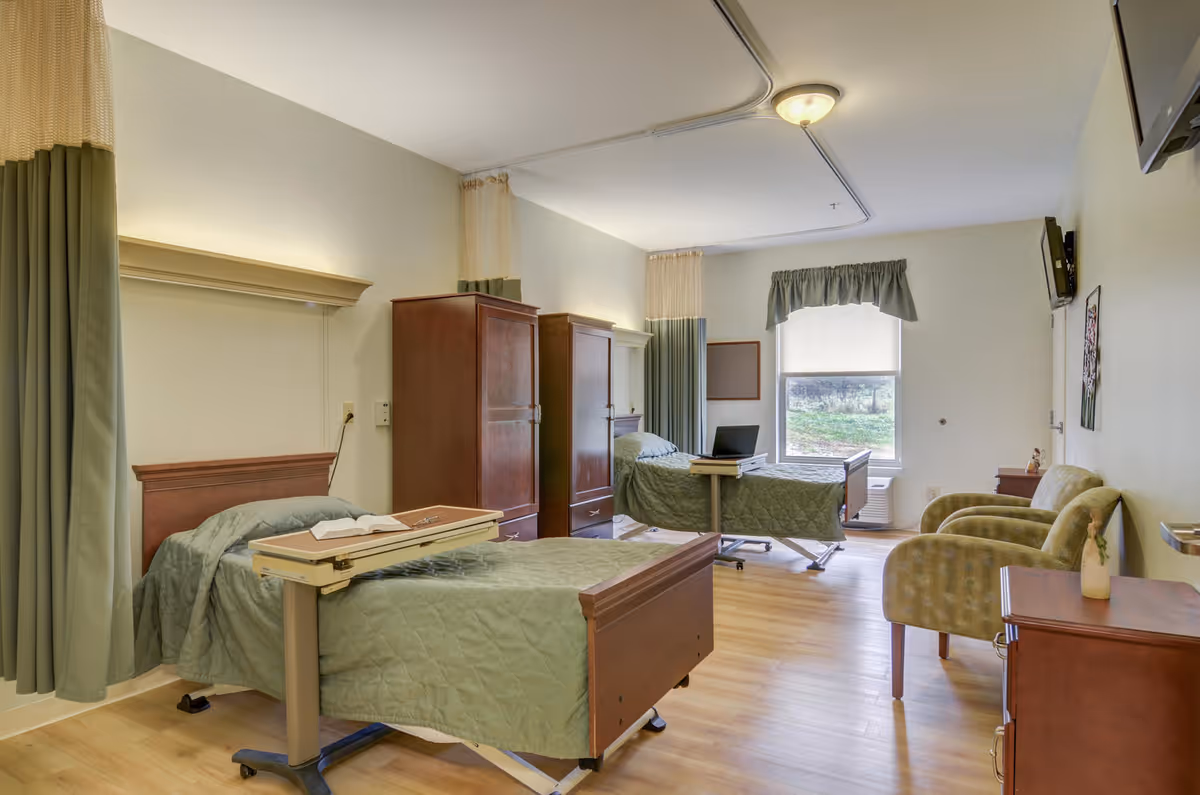 A bright and clean shared bedroom in a healthcare facility with two hospital-style beds covered in green quilts, two wooden wardrobes, two green armchairs, a small wooden dresser, a window with a green valance, and a wall-mounted TV.