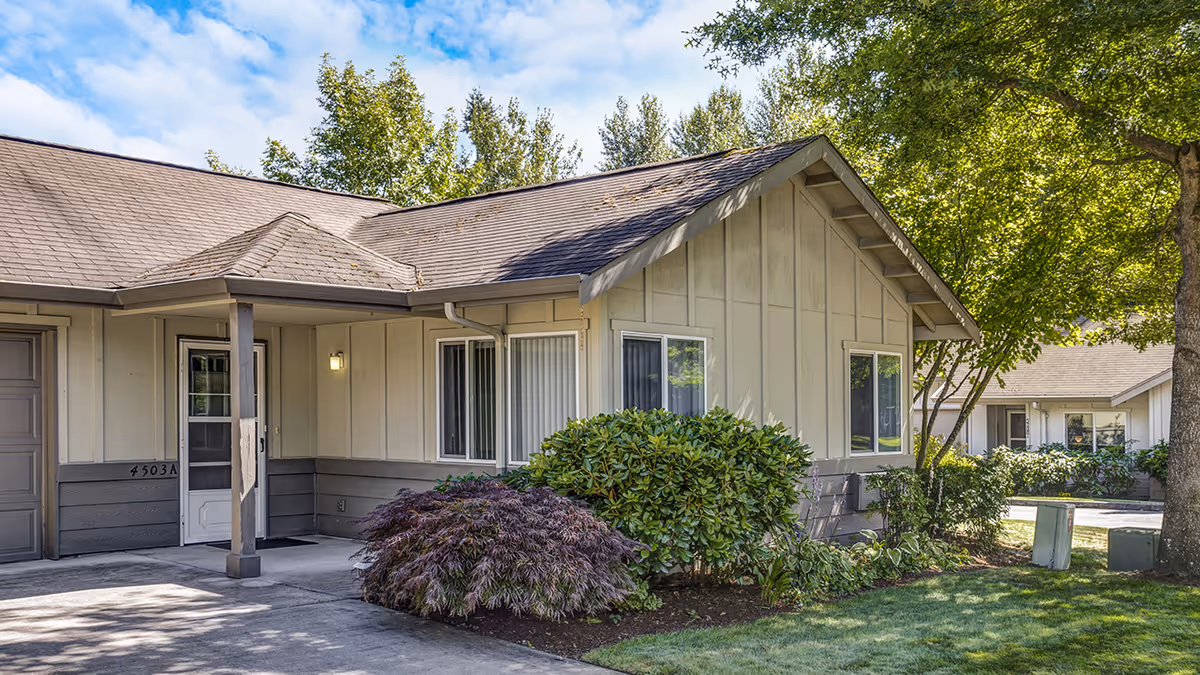 Exterior front of a single-story residential building with a covered entry, garage door, shrubs, and trees.