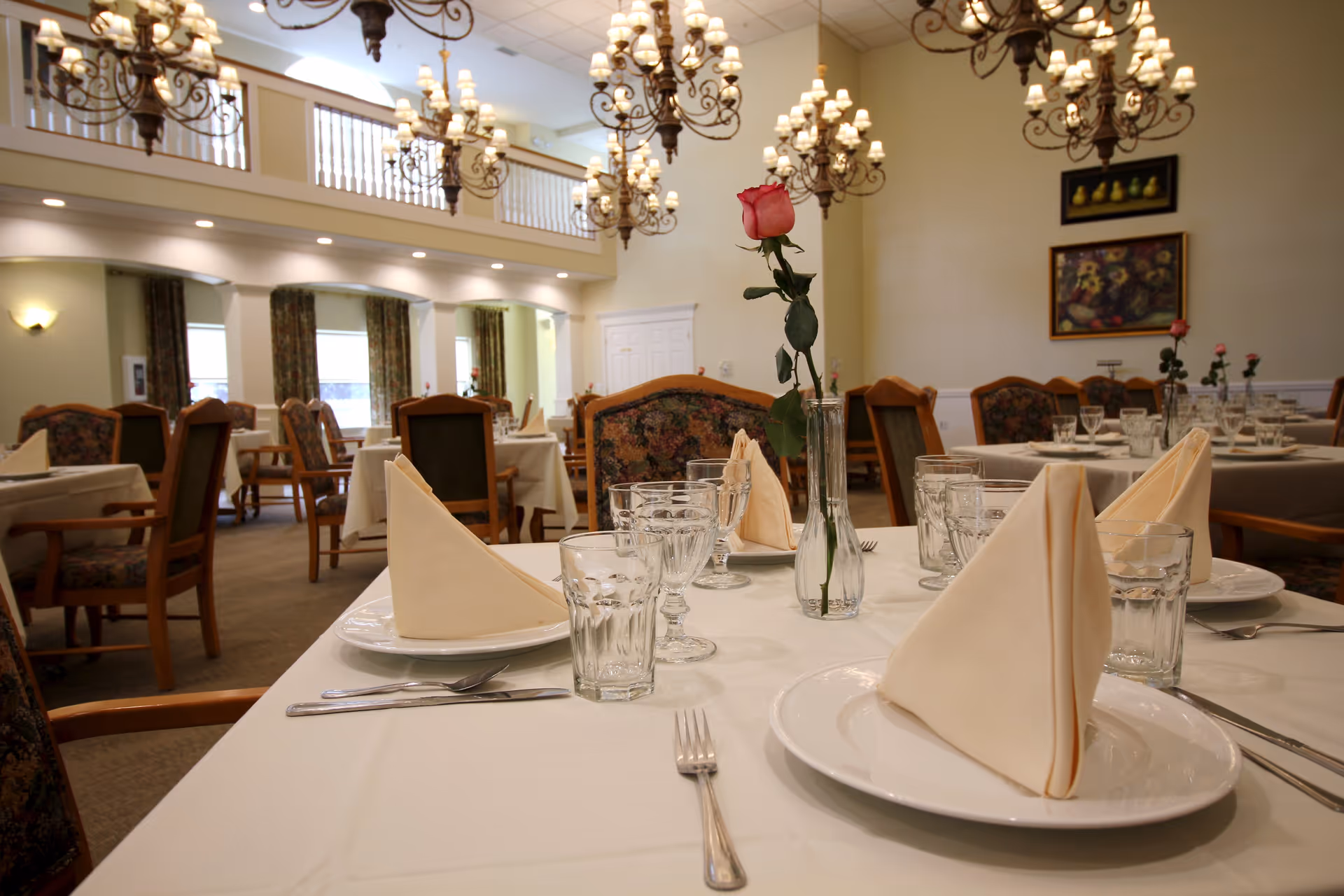 A well-lit dining room with tables set for a meal, featuring white tablecloths, folded beige napkins, glassware, and single pink roses in vases. The room has chandeliers hanging from the ceiling, floral upholstered chairs, and framed artwork on the walls.