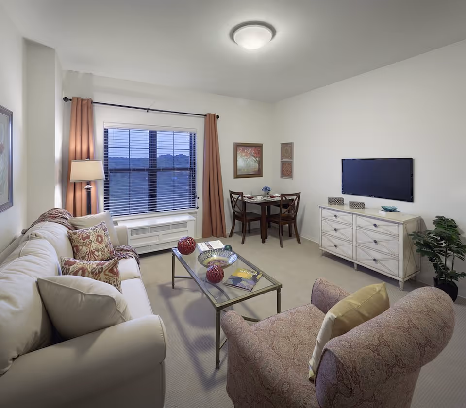 A cozy living room in a senior living facility featuring a beige sofa with patterned cushions, a glass coffee table with decorative items, a patterned armchair, a small dining table with two chairs, a wall-mounted flat screen TV above a white dresser, a floor lamp, and a window with brown curtains and blinds.