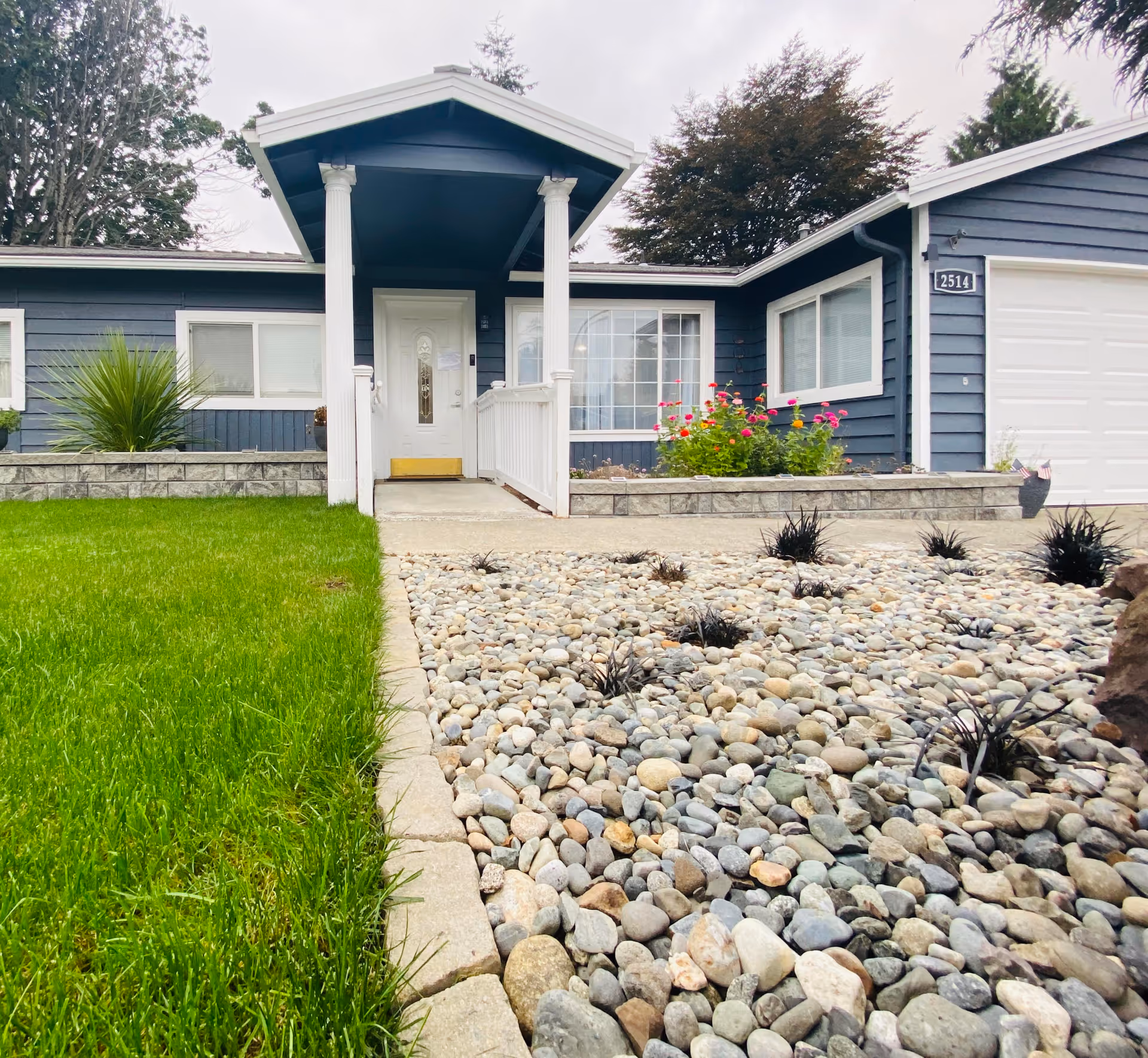 Front exterior of a blue single-story house with a covered white-pillared porch, green lawn and rock landscaping.