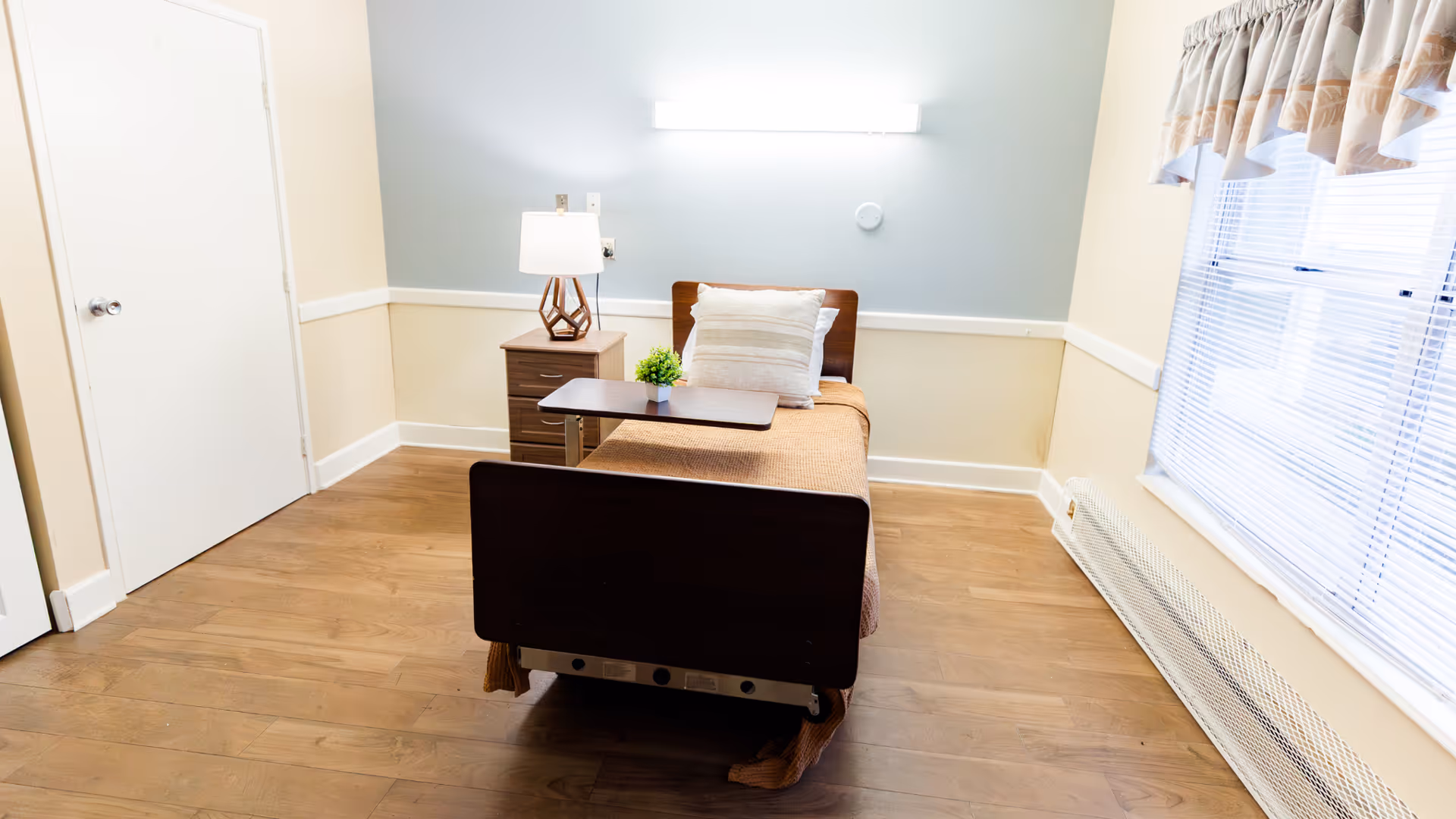 A simple, clean bedroom with a single hospital-style bed covered with a brown blanket and a pillow. Next to the bed is a wooden nightstand with a modern lamp and a small green plant. The room has light-colored walls, a large window with blinds and a valance, and wooden flooring.