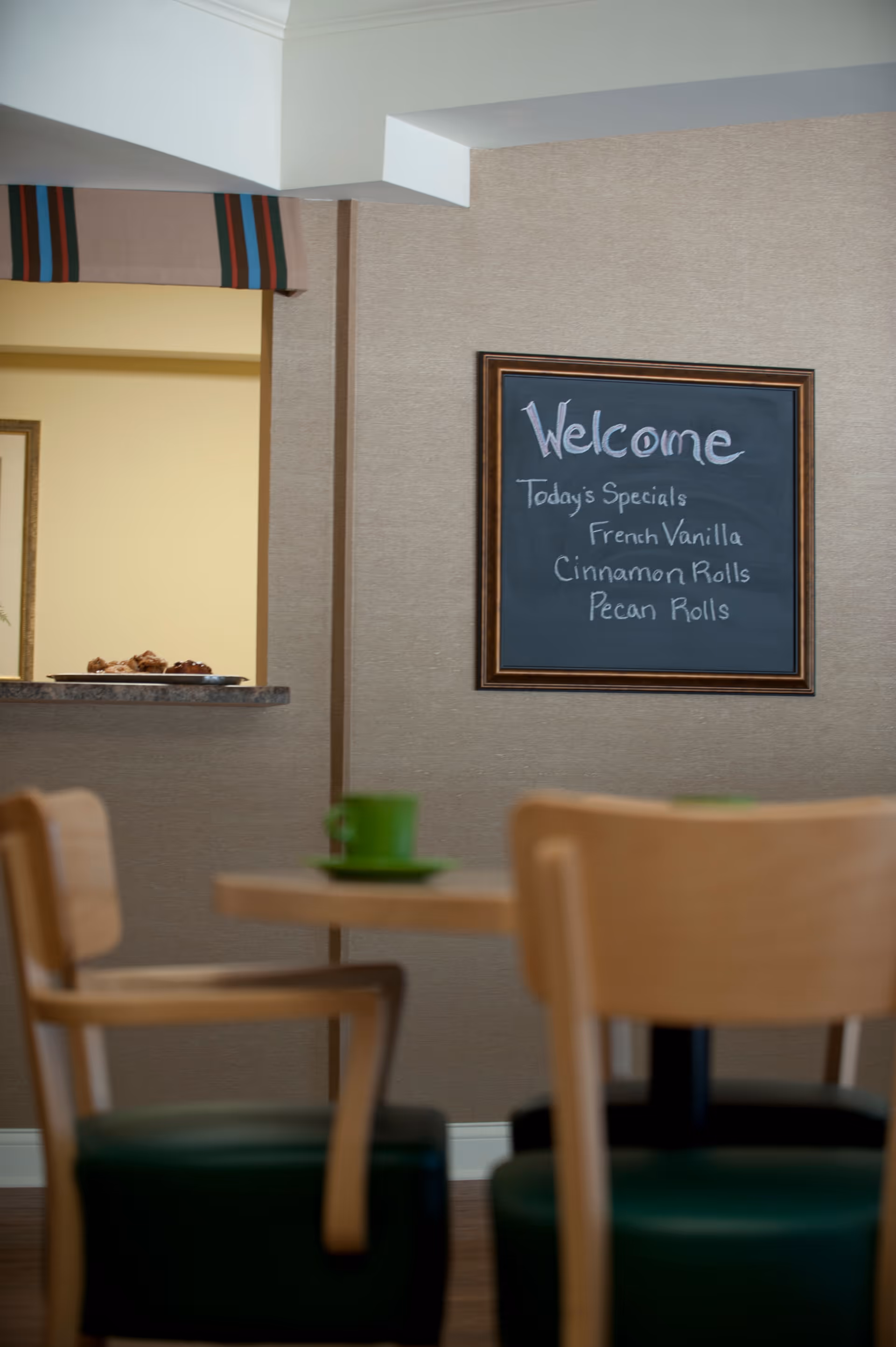 Small dining area with wooden chairs and a table in front of a wall-mounted chalkboard that says "Welcome" and lists pastry specials.