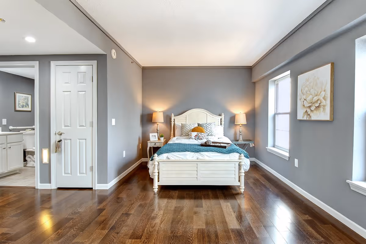 A bright bedroom with gray walls and wooden flooring featuring a white wooden bed with a blue throw blanket and multiple pillows. Two bedside tables with lamps flank the bed. A window on the right side lets in natural light, and a floral painting hangs on the wall. To the left, an open door reveals a bathroom with white cabinetry and a toilet.