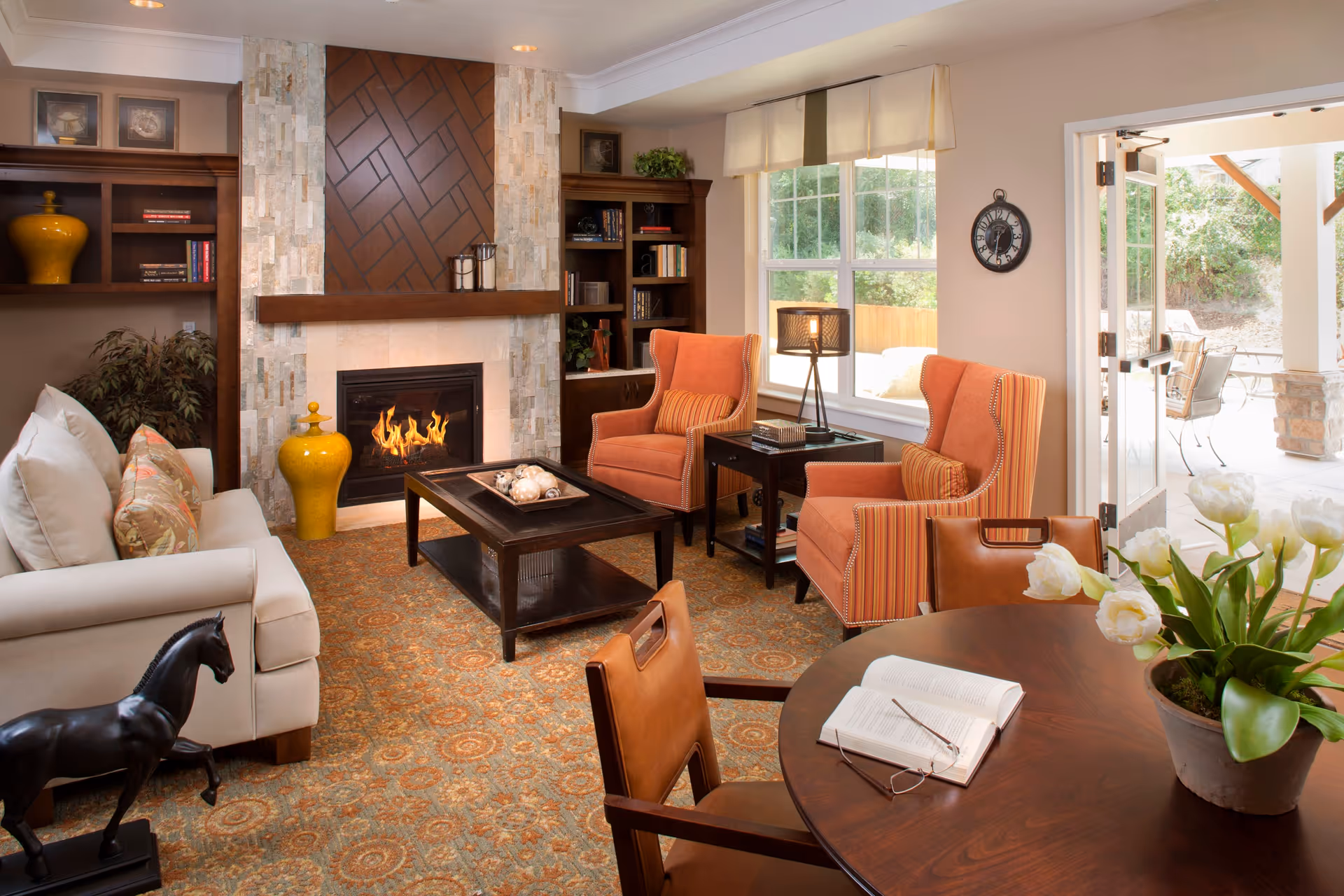 A cozy living room with a lit fireplace surrounded by stone and wood paneling. The room features a beige sofa with patterned cushions, two orange armchairs with striped cushions, a dark wood coffee table, and a round wooden table with a potted plant and an open book with reading glasses. There are built-in wooden shelves with books and decorative items, a window with a view of greenery outside, and a door leading to an outdoor patio area.