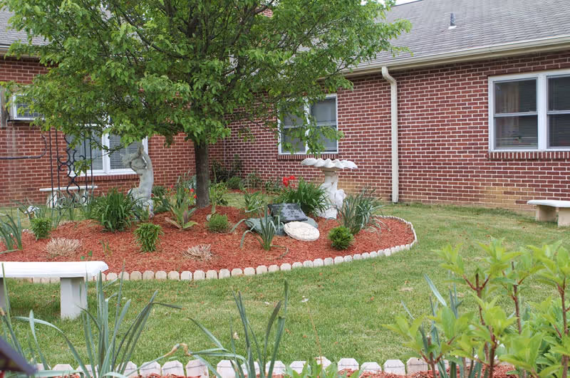 A landscaped garden area with a tree, various plants, and decorative elements including a birdbath and a small statue, bordered by a curved edging of bricks. The garden is situated next to a red brick building with several windows and a white bench on the grass nearby.