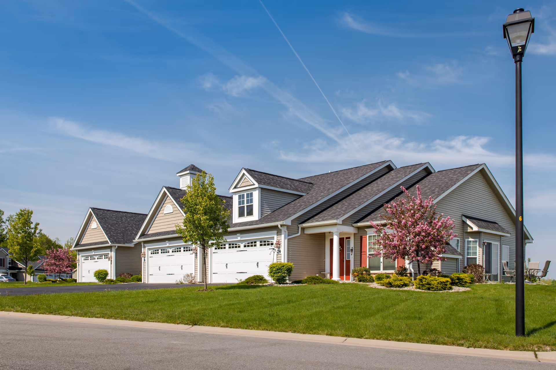 Exterior view of a single-story residential building with multiple attached garages, manicured green lawn, blooming pink trees, and a street lamp under a clear blue sky.