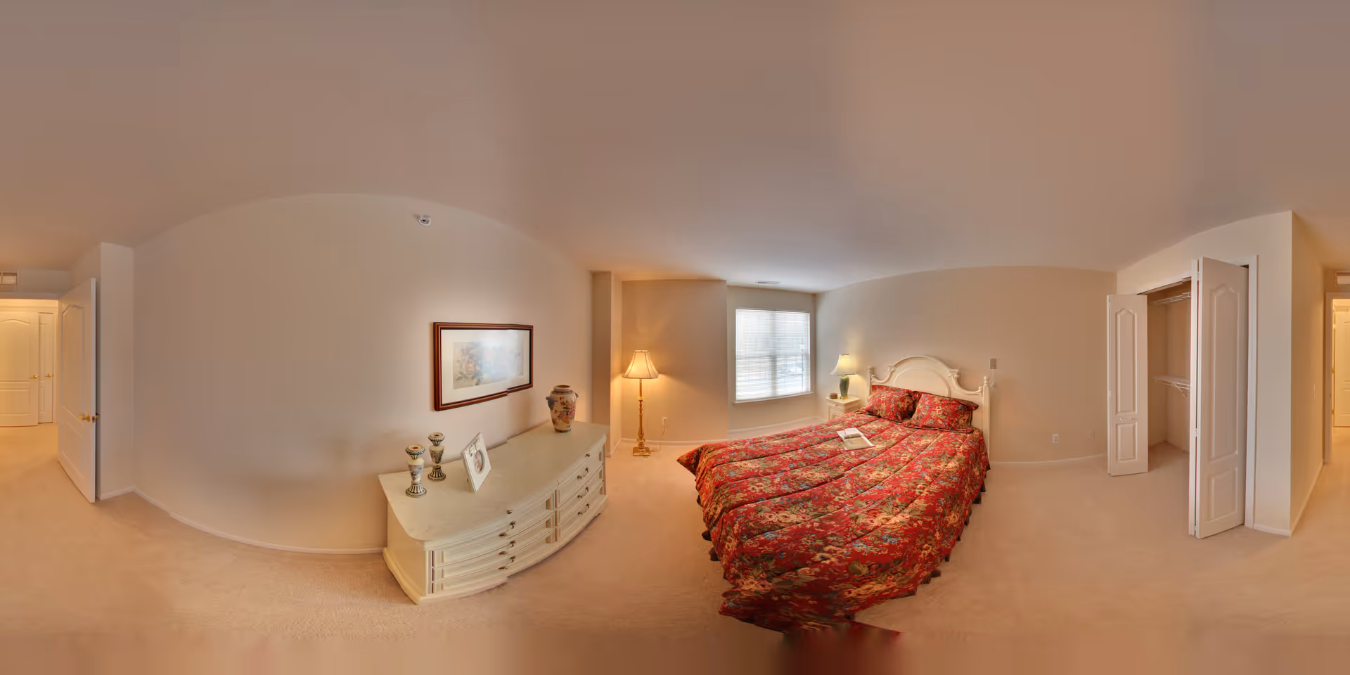 A cozy bedroom with beige walls and carpet, featuring a bed with a red floral bedspread, a white dresser with decorative items and framed artwork above it, a floor lamp, a bedside table with a lamp, a window with blinds, and an open closet with double doors.
