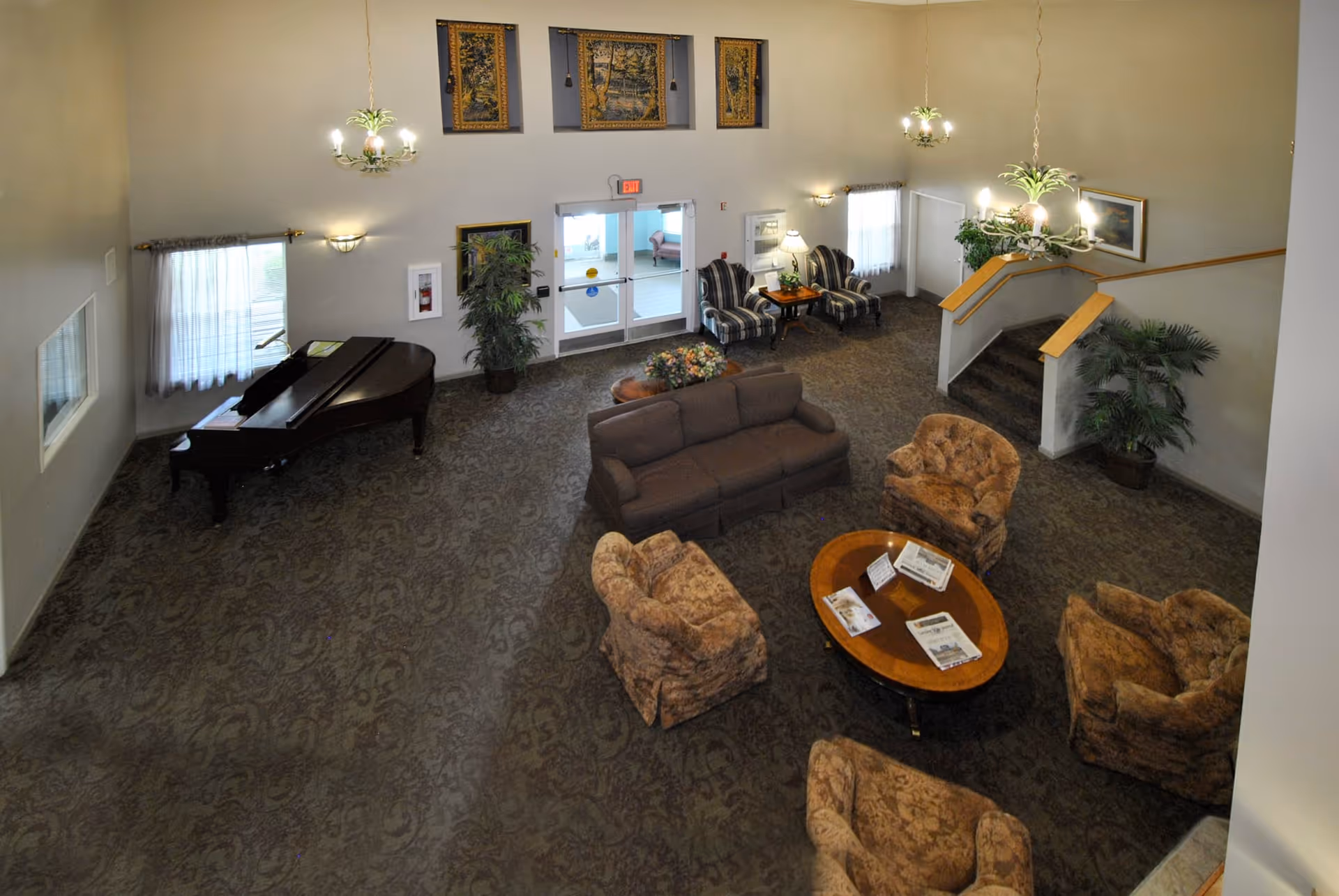A spacious living room area with patterned carpet, a brown sofa, four upholstered armchairs arranged around a wooden coffee table with magazines, two striped armchairs near a side table with a lamp, a grand piano near a window with sheer curtains, potted plants, wall sconces, hanging chandeliers, and three framed tapestries above a double glass door exit.