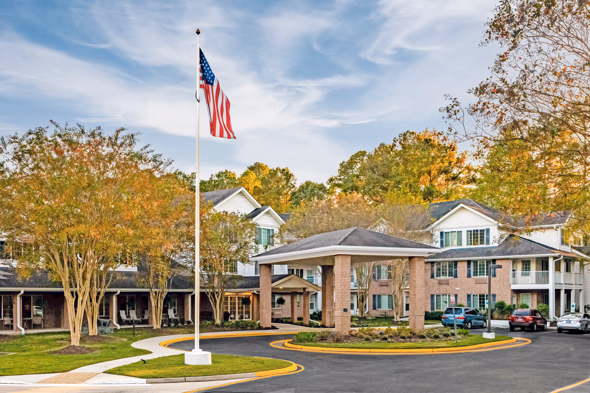 Exterior view of Lighthouse Pointe by Barclay House senior living facility with a circular driveway, an American flag on a flagpole, trees with autumn foliage, and several parked cars in front of a multi-story brick and siding building.