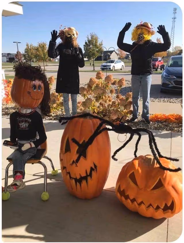 Outdoor Halloween decorations featuring three large pumpkin figures with faces and two standing figures wearing masks and wigs, set on a sidewalk with cars and trees in the background.