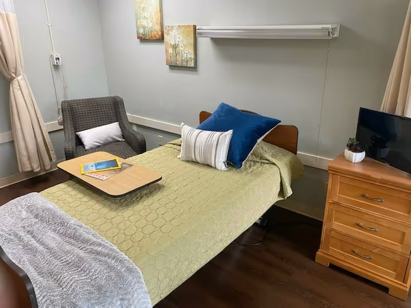 A small resident bedroom with a single bed covered by a green quilt and pillows, an armchair, overbed table, wooden dresser with a TV, and wall art.