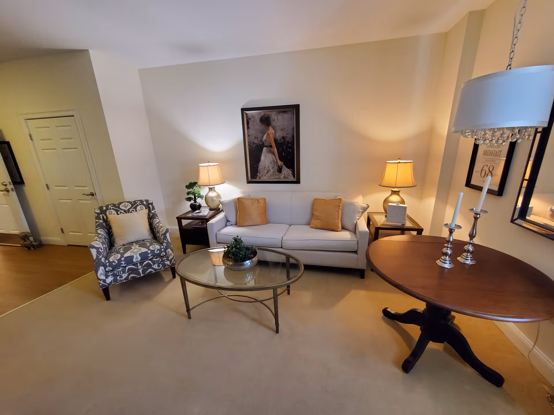 A cozy living room with a white sofa adorned with two beige pillows, a patterned armchair with a light cushion, a glass coffee table with a small plant centerpiece, two side tables each with a lamp, a round wooden table with two silver candlesticks, and a painting of a woman in a white dress on the wall.