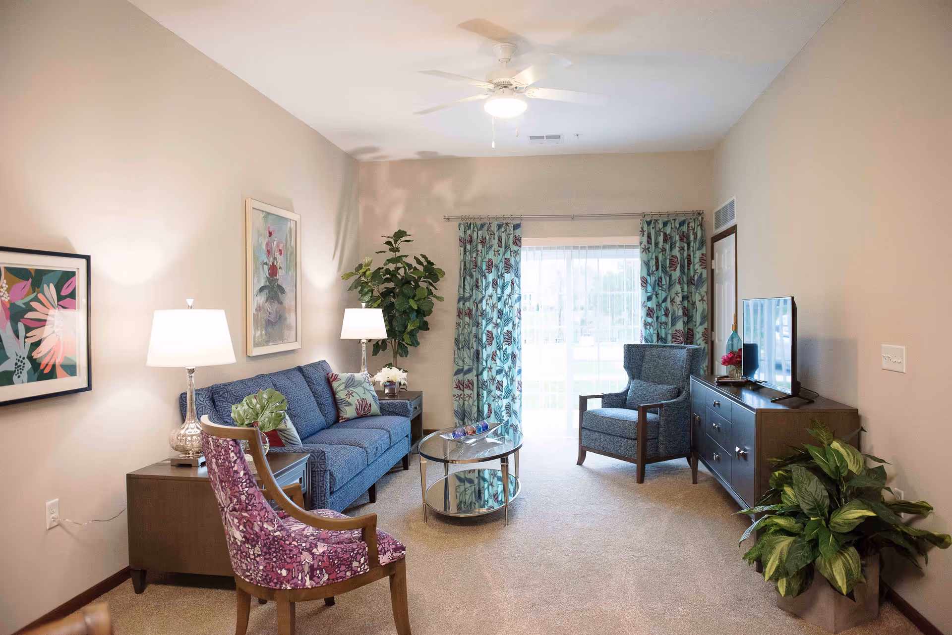 A cozy living room with a blue sofa, a purple patterned armchair, a gray armchair, two side tables with lamps, a glass coffee table, a TV on a wooden dresser, potted plants, floral curtains, and framed artwork on the walls.