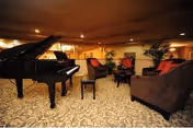 Warmly lit communal lounge with a grand piano, upholstered chairs and sofas arranged on patterned carpet.