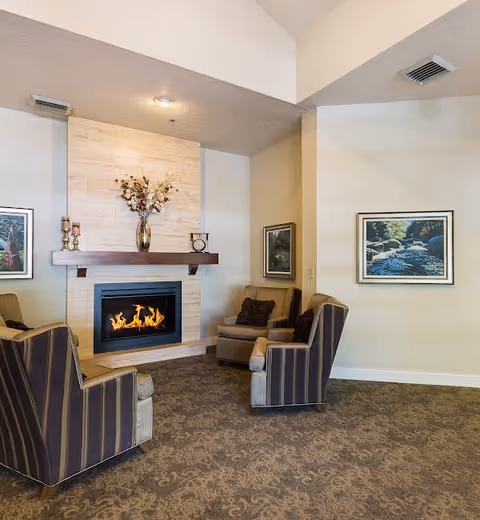 A cozy living room area with a lit fireplace set in a tiled wall. Above the fireplace is a wooden mantel decorated with a vase of dried flowers and two candle holders. Four upholstered armchairs with striped and solid patterns are arranged around the fireplace. The walls are adorned with framed landscape paintings, and the floor is covered with a patterned carpet.