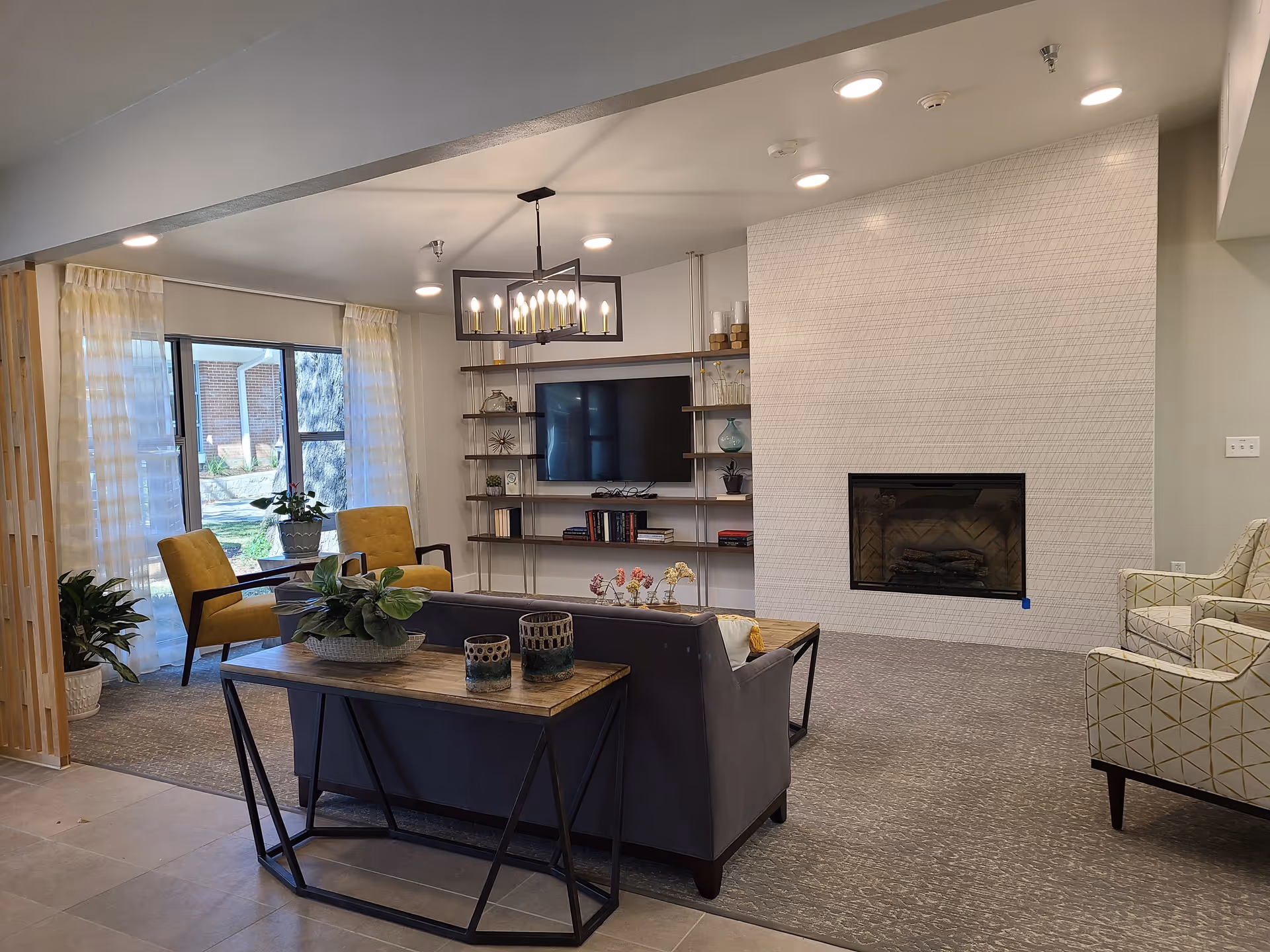 A modern living room with a gray sofa, two yellow armchairs, and two patterned white armchairs. There is a wooden console table behind the sofa with decorative items and plants. A flat-screen TV is mounted on the wall with shelves holding books and decor. The room features a fireplace with a textured white tile surround and large windows with sheer curtains letting in natural light.