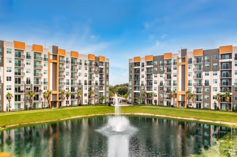 Two modern multi-story residential buildings facing each other with balconies, surrounding a pond with a water fountain in the center, under a clear blue sky.