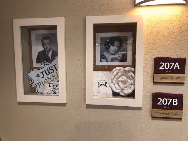 Two framed shadow boxes mounted on a beige wall, each containing a black and white portrait photo and decorative items. The left frame has a photo of a man, a sign that says 'Just Married,' and a small card with partial text. The right frame has a photo of a woman, a white decorative flower, and a card that says 'You are loved.' To the right of the frames are two maroon and gold room number plaques labeled 207A James Stewart and 207B Elizabeth Taylor. A wall light fixture is above the right frame.