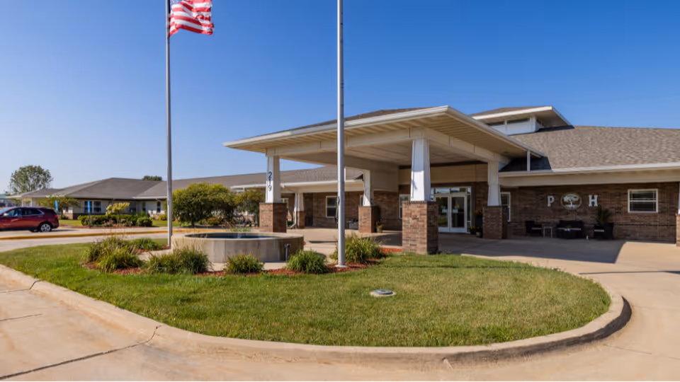 Front entrance of a single-story senior living facility with a covered porte-cochere, flagpole, and circular driveway.