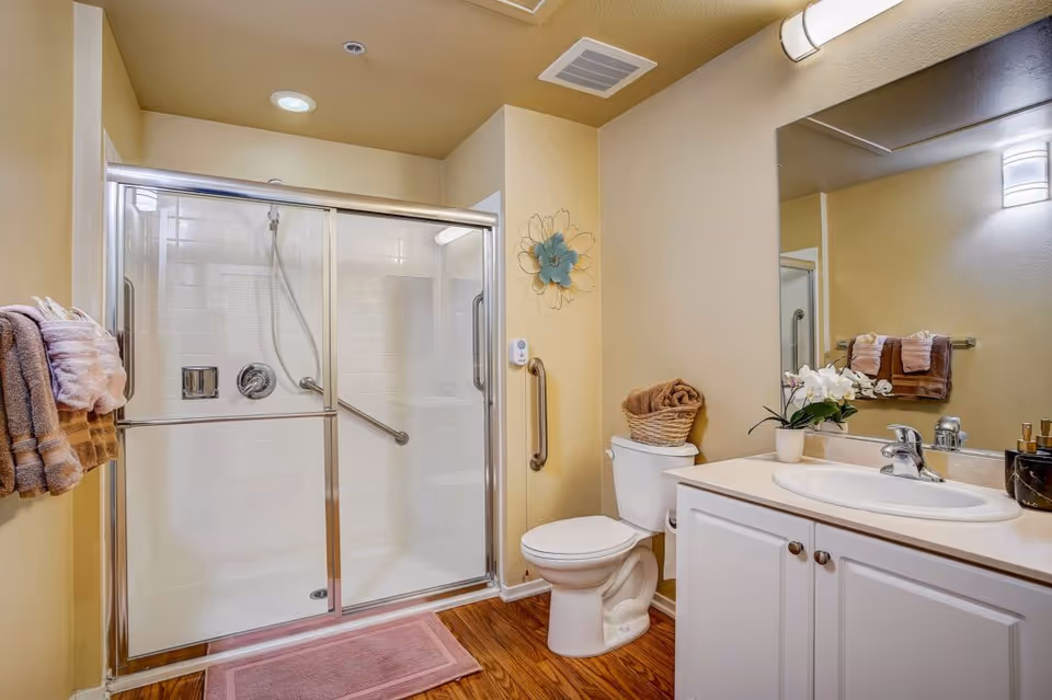 A clean, accessible bathroom with a glass-enclosed walk-in shower, toilet with grab bar, and a white vanity with mirror and decorative towels.