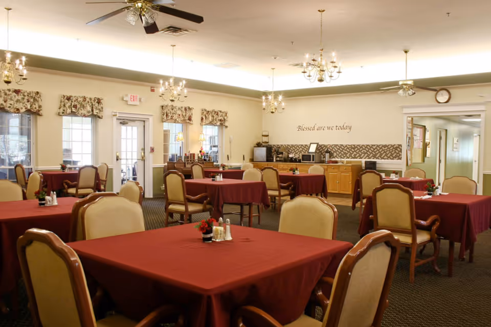 Dining room with tables covered in red tablecloths, upholstered chairs, chandeliers, and a service counter beneath a wall sign reading "Blessed are we today."