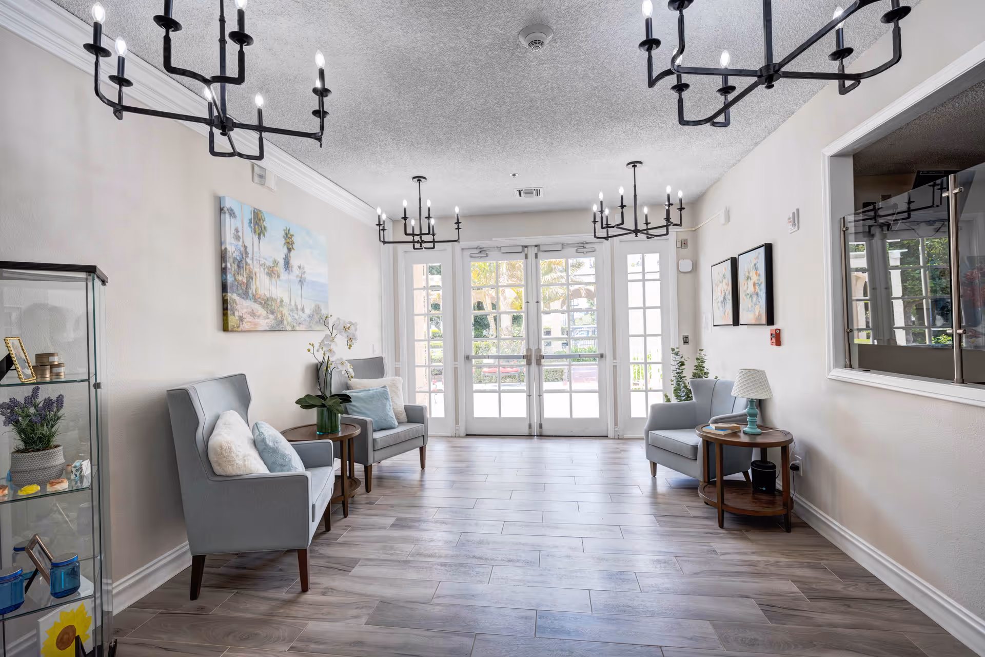 A bright and airy sitting area with light gray armchairs, small wooden side tables, and decorative pillows. The room features large glass double doors letting in natural light, modern black chandeliers hanging from the ceiling, a glass display cabinet with decorative items on the left, and framed artwork on the walls.