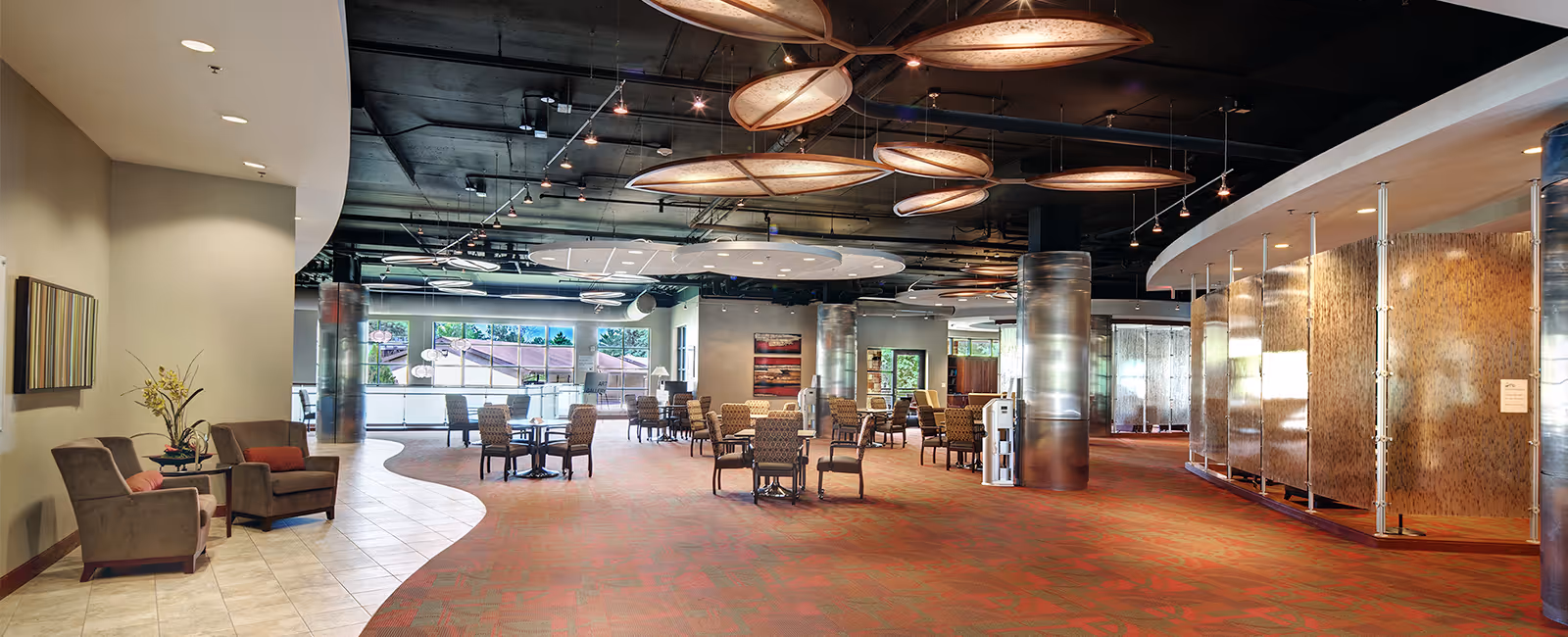 Spacious and modern common area with multiple seating arrangements including armchairs and tables, large windows letting in natural light, decorative ceiling panels, and metallic columns. The space has a mix of carpet and tile flooring and features artwork on the walls.