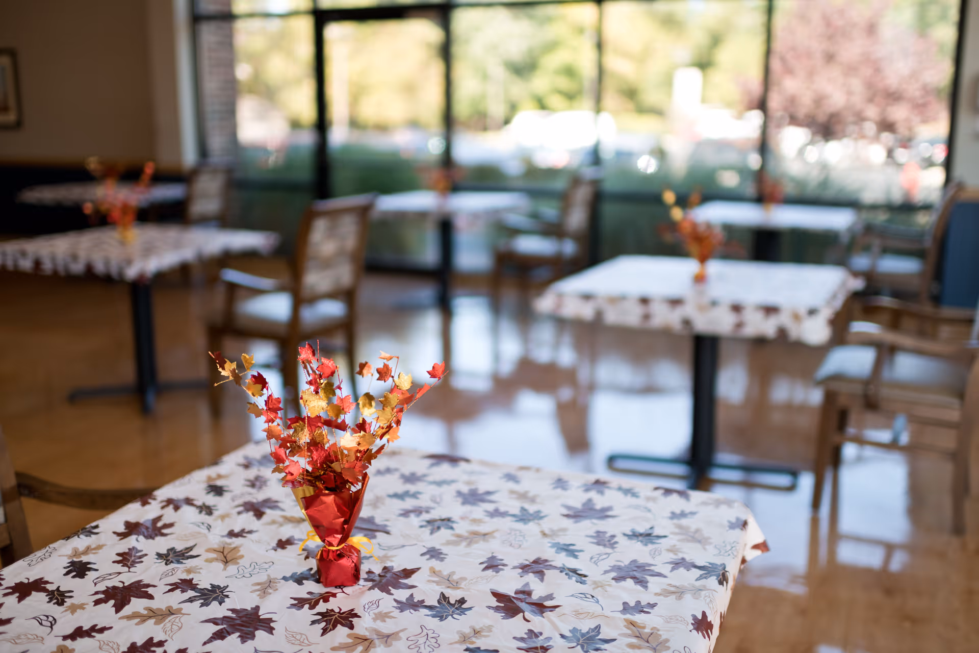 A dining area with several tables covered in white tablecloths decorated with autumn leaf patterns. Each table has a small centerpiece featuring red and yellow artificial autumn leaves. The room has large windows letting in natural light and wooden chairs around the tables.