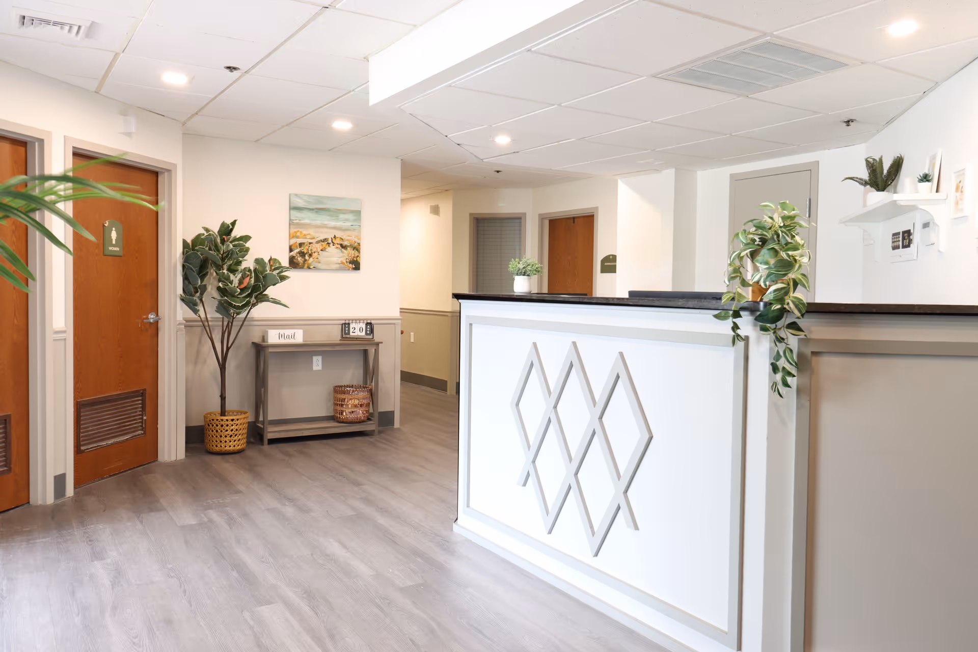 Bright and clean reception area with a white front desk featuring diamond-shaped wall decor. To the left, there are two wooden doors, one marked as a restroom. A small table with a plant and decorative items is against the wall, along with a large potted plant. The floor is light gray wood, and the walls are white with beige paneling. Ceiling lights illuminate the space.