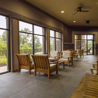 A spacious indoor seating area with wooden chairs and tables arranged near large windows that provide a view of greenery outside. The room has tiled floors, ceiling fans, and a stone accent wall near the entrance to an outdoor patio.