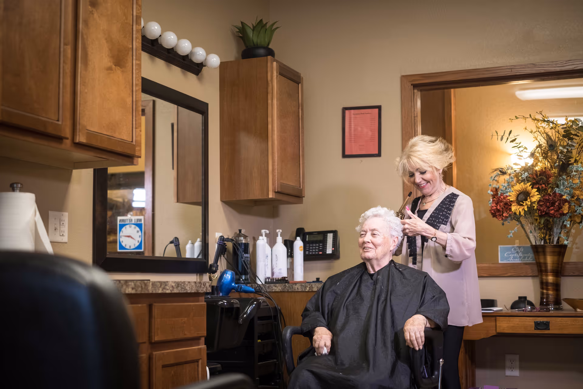 An elderly woman with white hair is sitting in a salon chair wearing a black cape while a hairstylist with blonde hair is cutting her hair. The room has wooden cabinets, a large mirror, hair care products on the counter, and a vase with colorful flowers in the background.