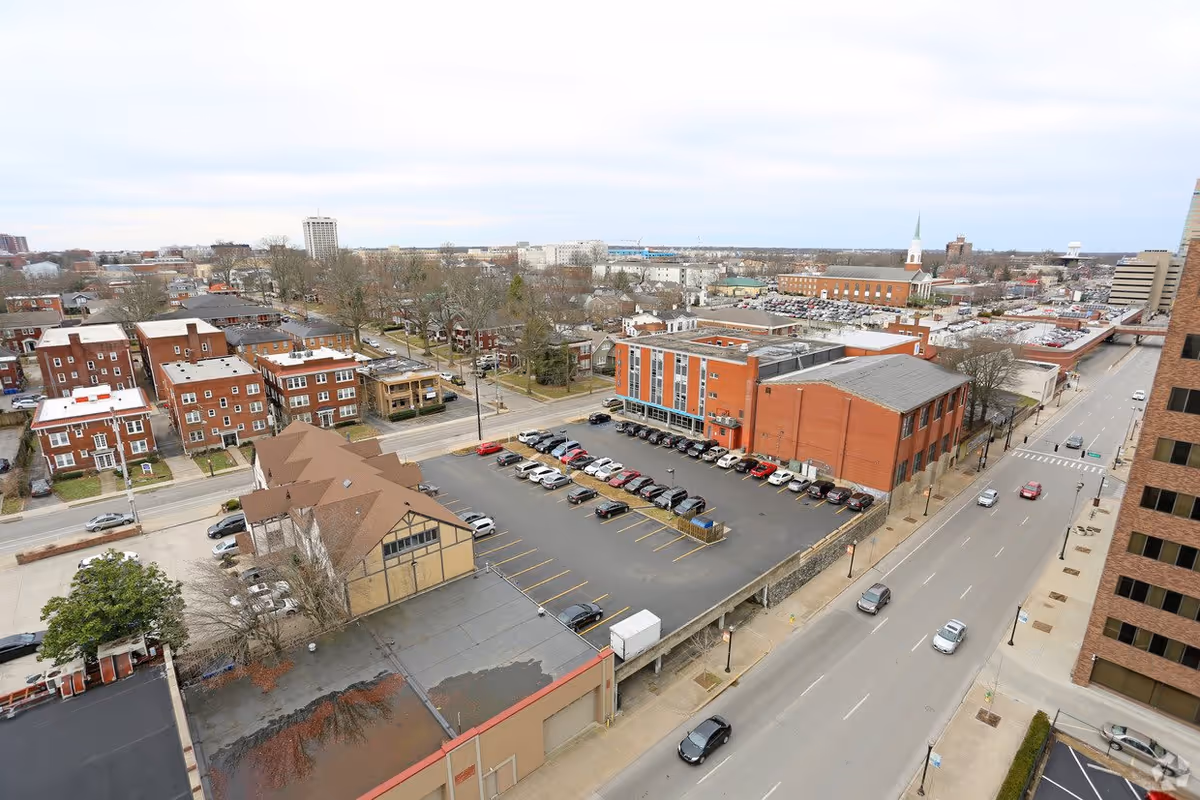 Aerial view of an urban area showing multiple buildings, a parking lot with cars, streets with vehicles, and a distant cityscape under a cloudy sky.