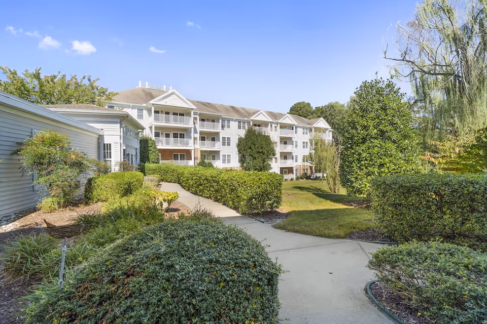 A paved walkway surrounded by well-maintained bushes and greenery leading to a multi-story residential building with balconies under a clear blue sky.