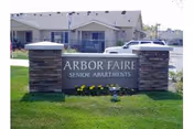 Stone sign with the text 'Arbor Faire Senior Apartments' in front of a residential building with parked cars and a well-maintained lawn.