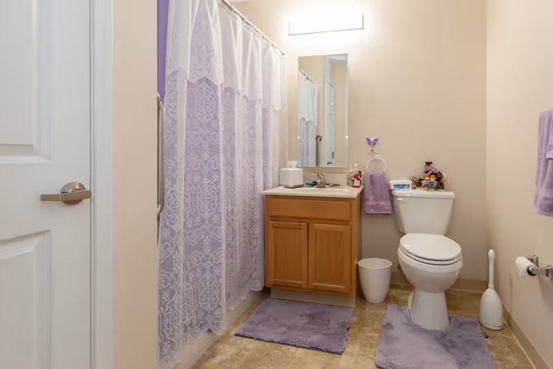 A clean bathroom with beige walls and tiled floor. There is a white door on the left, a shower with a purple patterned curtain, a wooden vanity with a sink and mirror above it, a white toilet with a floral decoration on the tank, and purple bath mats on the floor. Purple towels hang on the wall and a white trash bin and toilet brush are beside the toilet.