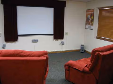 Red upholstered chairs face a white projection screen in a small lounge area with burgundy curtains.