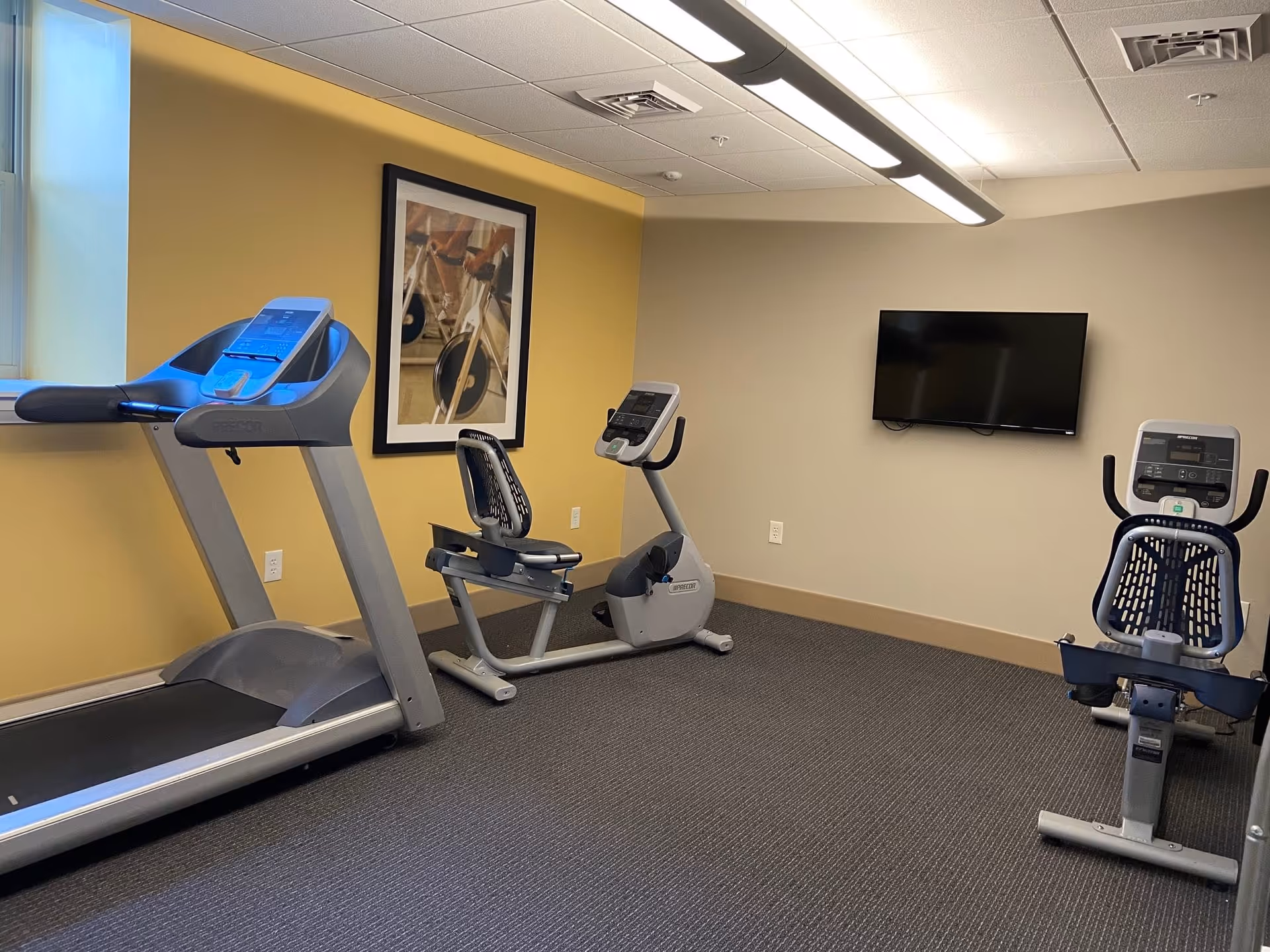 A small fitness room with a treadmill, two recumbent exercise bikes, a wall-mounted flat screen TV, and a framed picture of a stationary bike on a yellow accent wall.